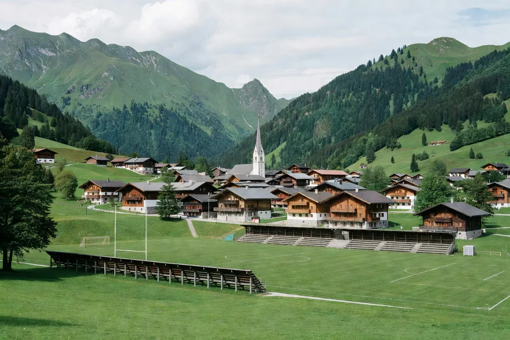 Austrian alpine valley town with green mountains, a church spire and a small football stadium below