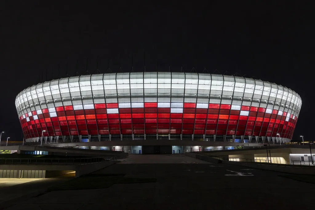 Polish national football stadium with red and white exterior cladding glowing under night lighting, wide exterior view