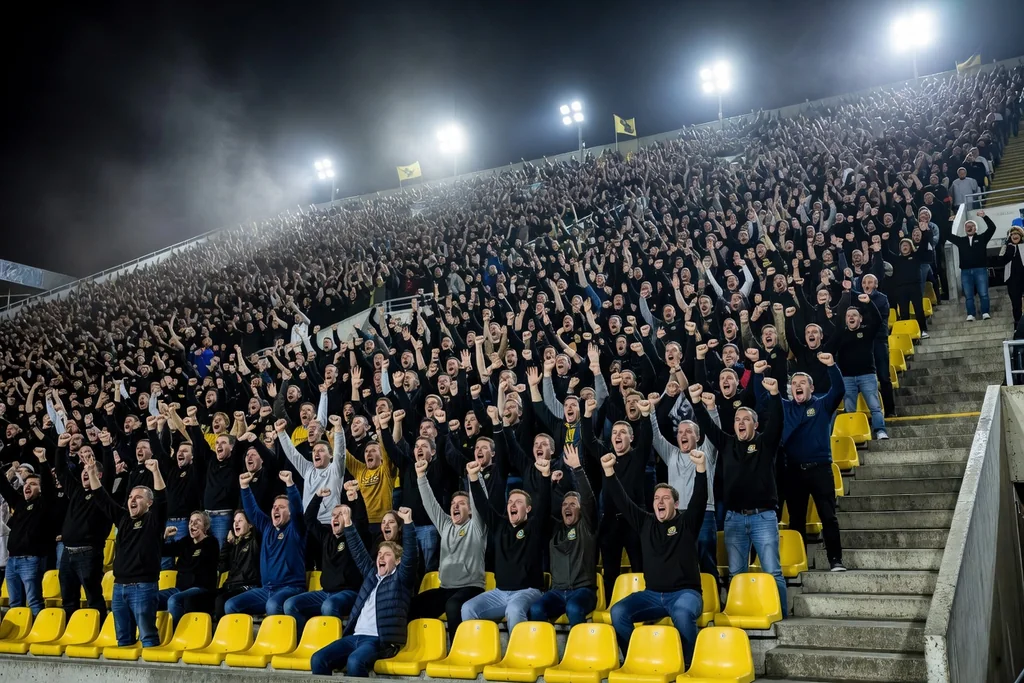 A huge steep football terrace at night packed with supporters seen from pitch level: yellow seats